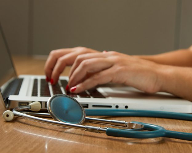 a doctors hands typing on a laptop with a stethoscope next to the keyboard
